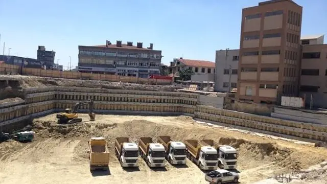 Binalar arasında, çevresi iksa duvarlarıyla desteklenen derin kazı alanında ekskavatör ve kamyonlar
